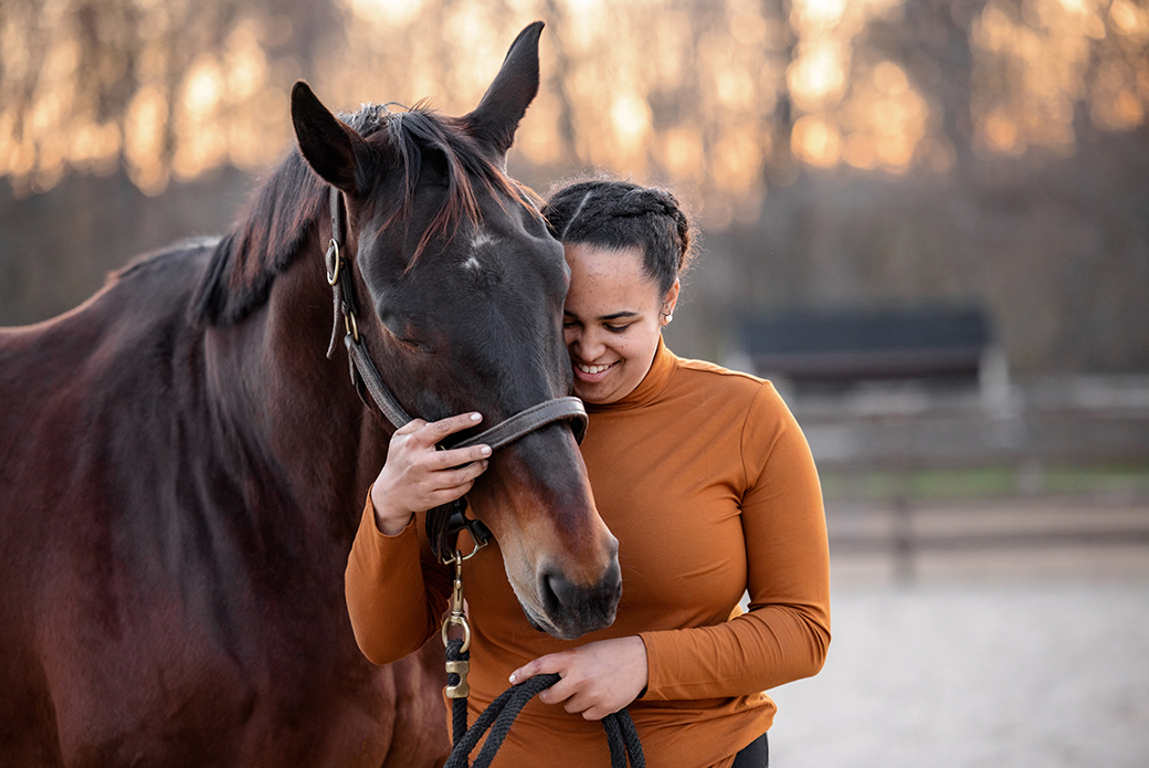 A woman in an orange long sleeve shirt hugging a dark brown horse's head