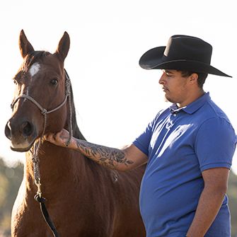 a man with a black cowboy hat blue shirt and tattoo on his arm scratching the cheek of a brown horse with a white forehead mark
