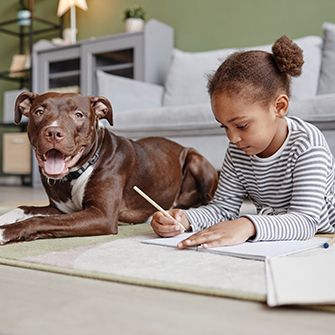 A little girl in a stripe shirt writing in a notebook on the floor next to large brown dog